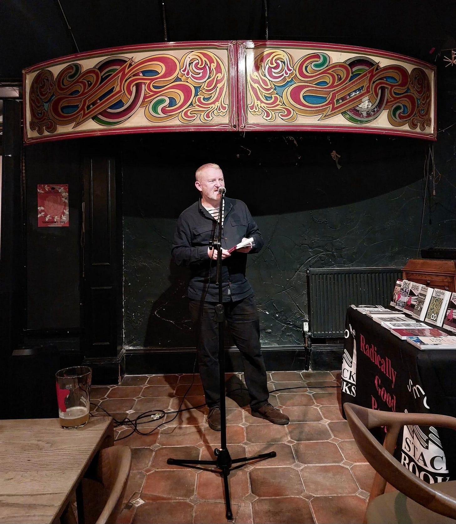 Greying-ginger man dressed in black/dark blue with book of poems in his hand, framed by an old fairground hoarding.