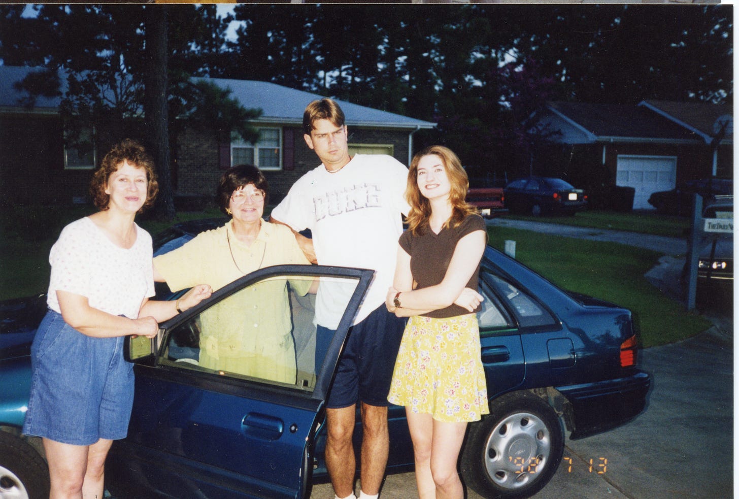 Four white people, two older women, a tall young man, and a young woman next to a car in the suburbs. Date on photo is July 13, 1998.