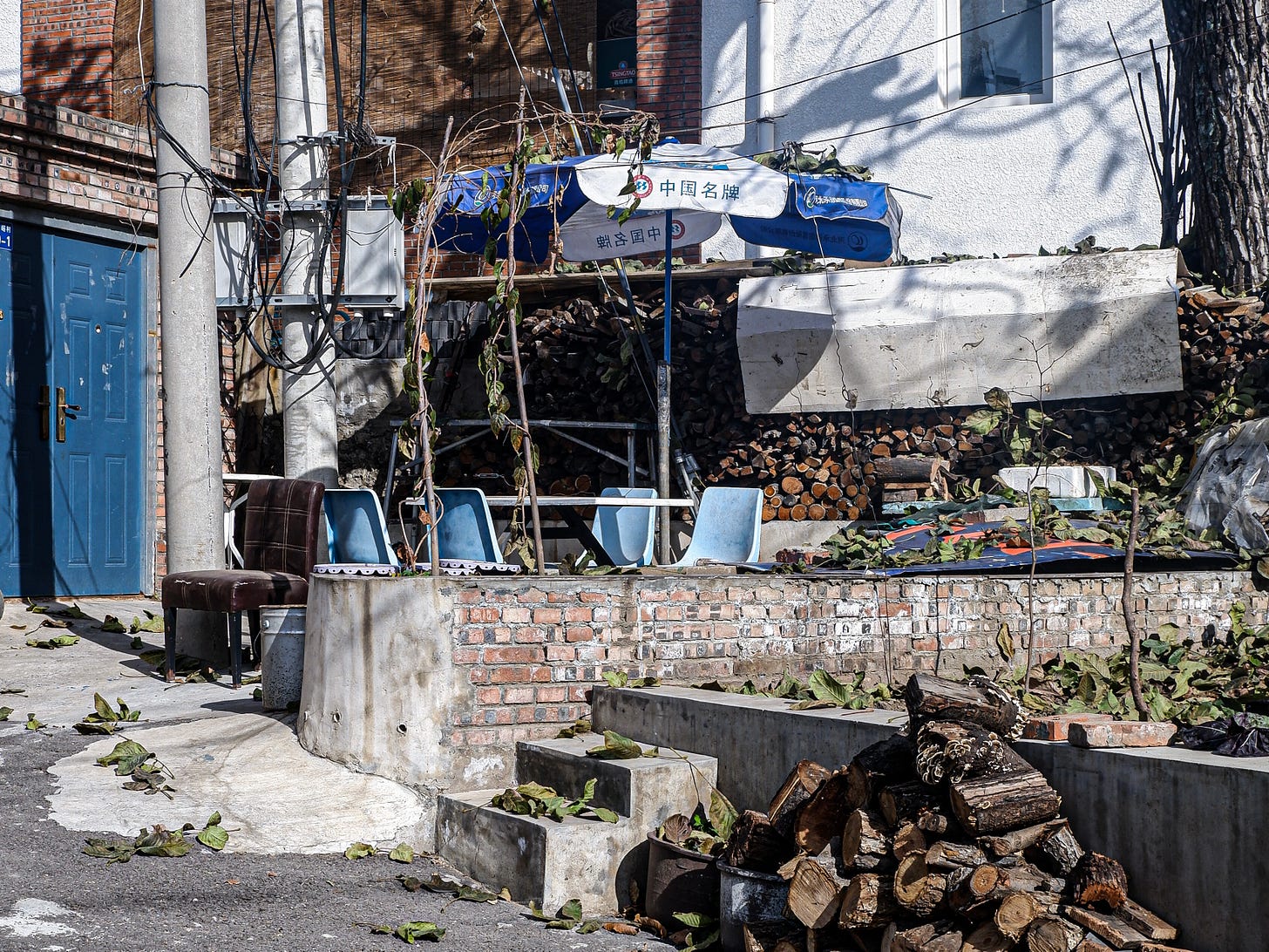 Neatly stacked firewood sits beside a worn leather armchair and a patio umbrella parked outside a vibrant blue door