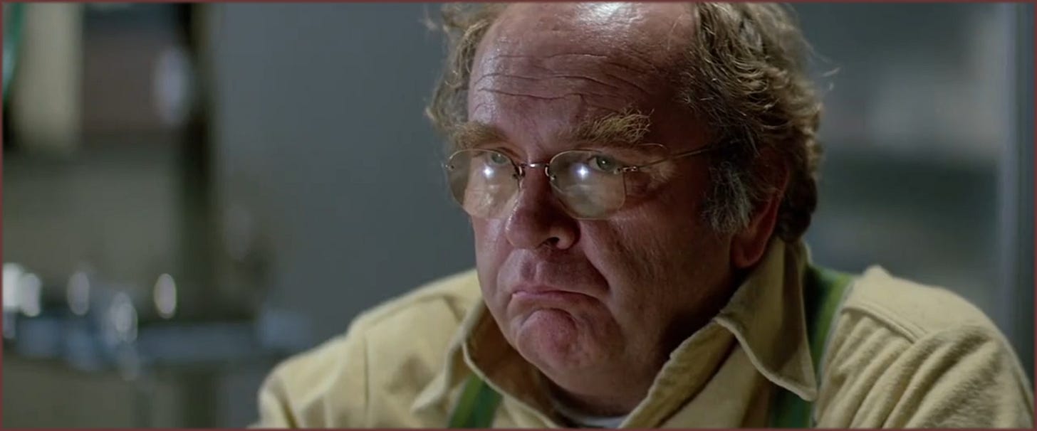 Close-up from The Thing (1982): Blair (Wilford Brimley) staring at a computer screen, glasses reflecting the light, expression tense and inward. The moment before the simulation results destroy his composure.