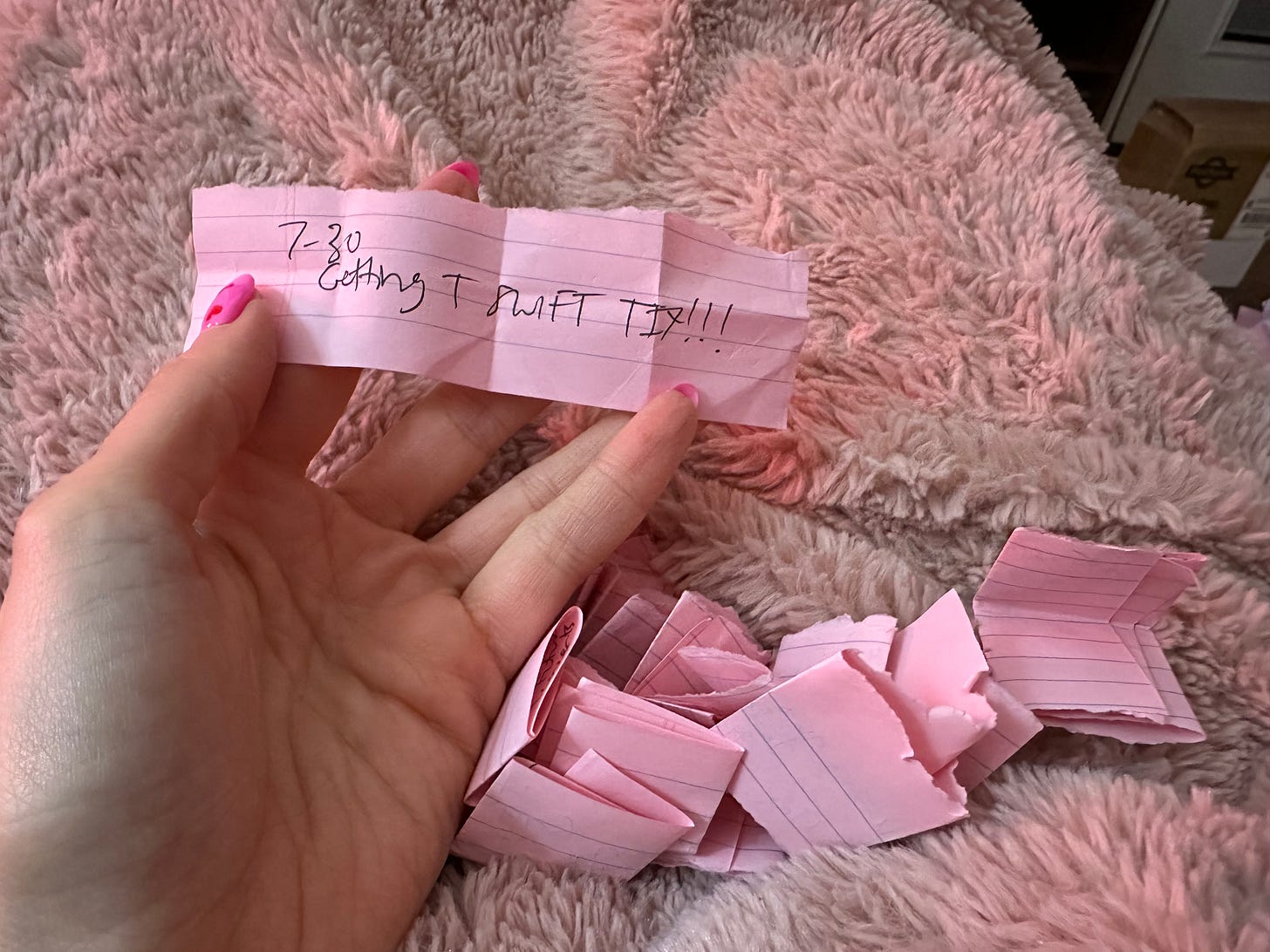 A woman's left hand holds up a pink scrap of paper with "7/30 Getting T SWIFT TIX!!!" written on it