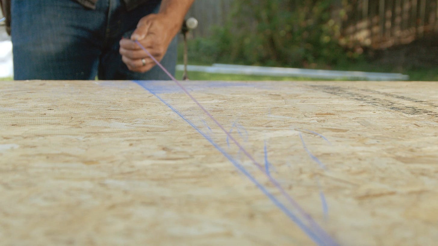 A carpenter snaps a blue chalk line on a piece of chipboard sheathing