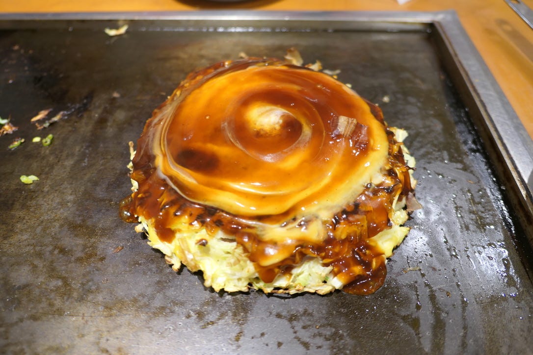An okonomiyaki on a rectangular griddle at a restaurant. A spiral of brown sauce is on top of the pancake, with cabbage showing on the side.. An okonomiyaki on a rectangular griddle at a restaurant. A spiral of brown sauce is on top of the pancake, with cabbage showing on the side..