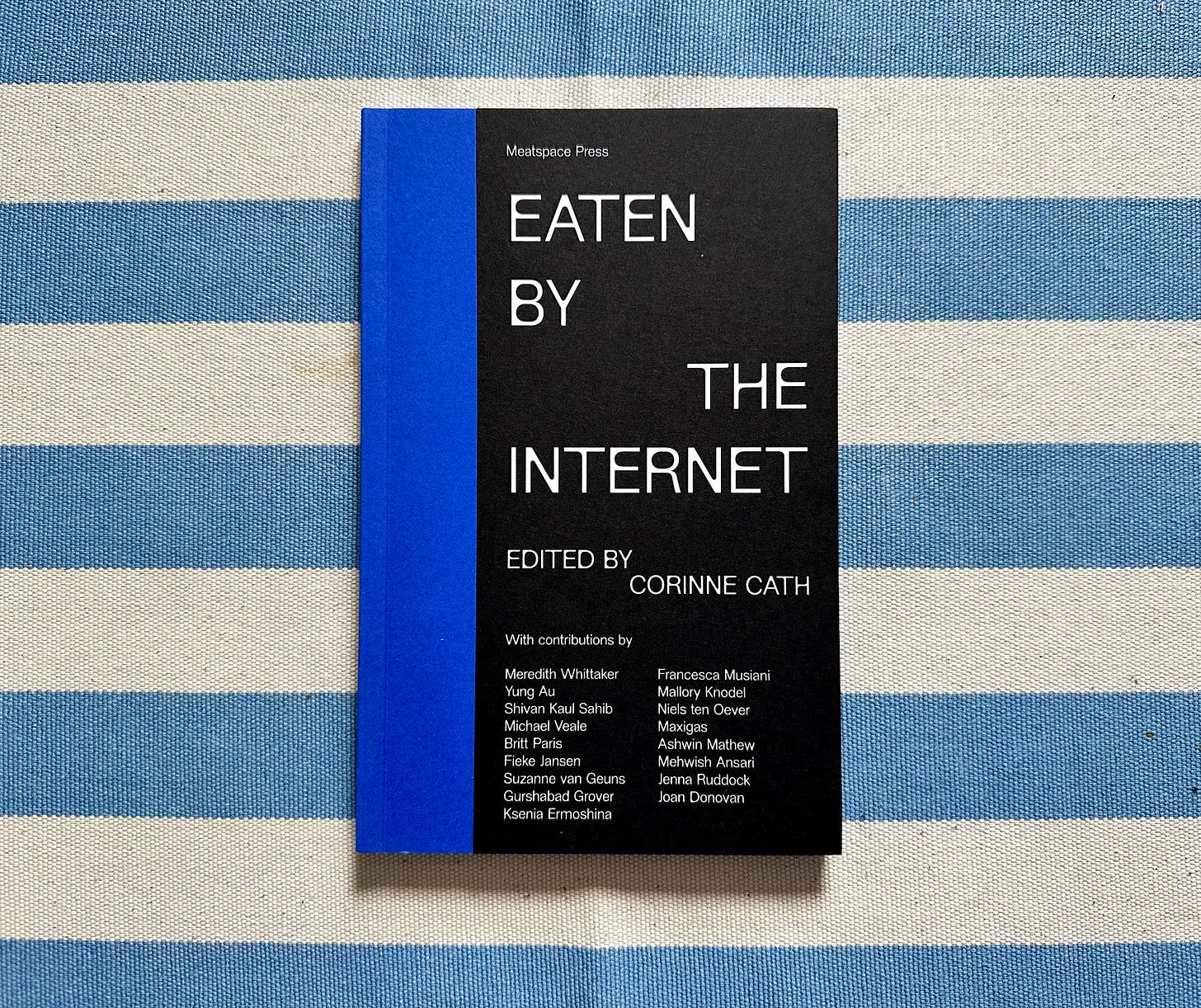 Picture of a book titled “Eaten by the Internet” edited by Corinne Cath on a textured cotton tablecloth with thick off-white and light blue stripes. The cover is vertically bisected half blue and half black, with the title and the names of its contributors on the cover.