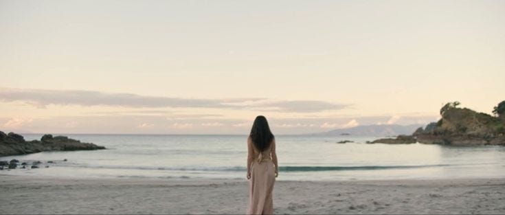 This may contain: a woman standing on top of a sandy beach next to the ocean in front of an island