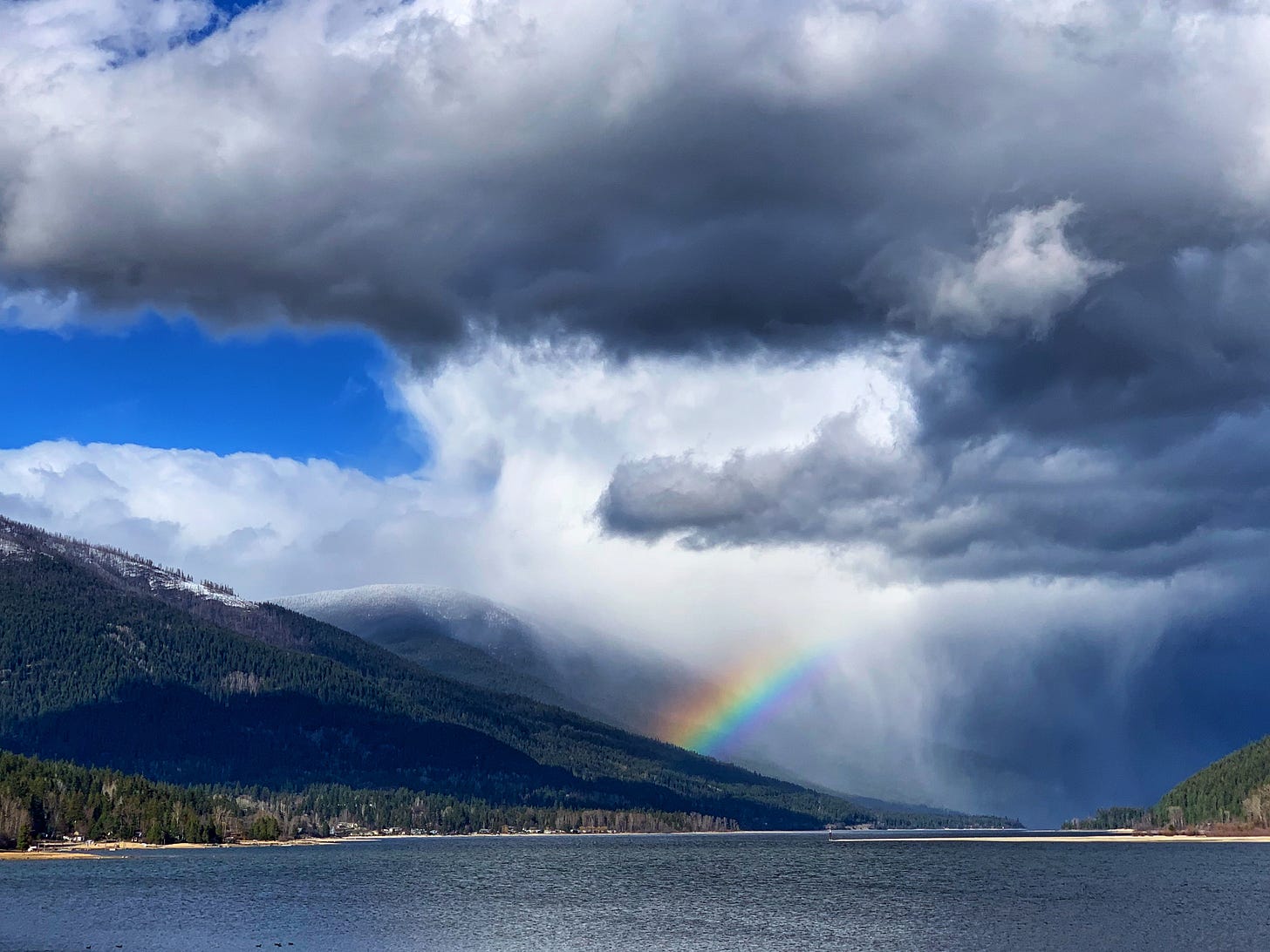Kootenay Lake in Nelson, British Columbia, with forested mountains, dramatic clouds, and a vibrant rainbow over the water