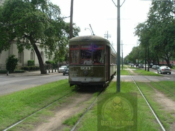  This is a photograph of a historic green streetcar, number 933, sitting on a grassy median between its tracks. It appears to be on St. Charles Avenue in New Orleans, with a street and large trees lining both sides. The streetcar is facing the viewer and is stopped. In the background, there are other cars on the road and a large white building to the left. The sky is slightly overcast.