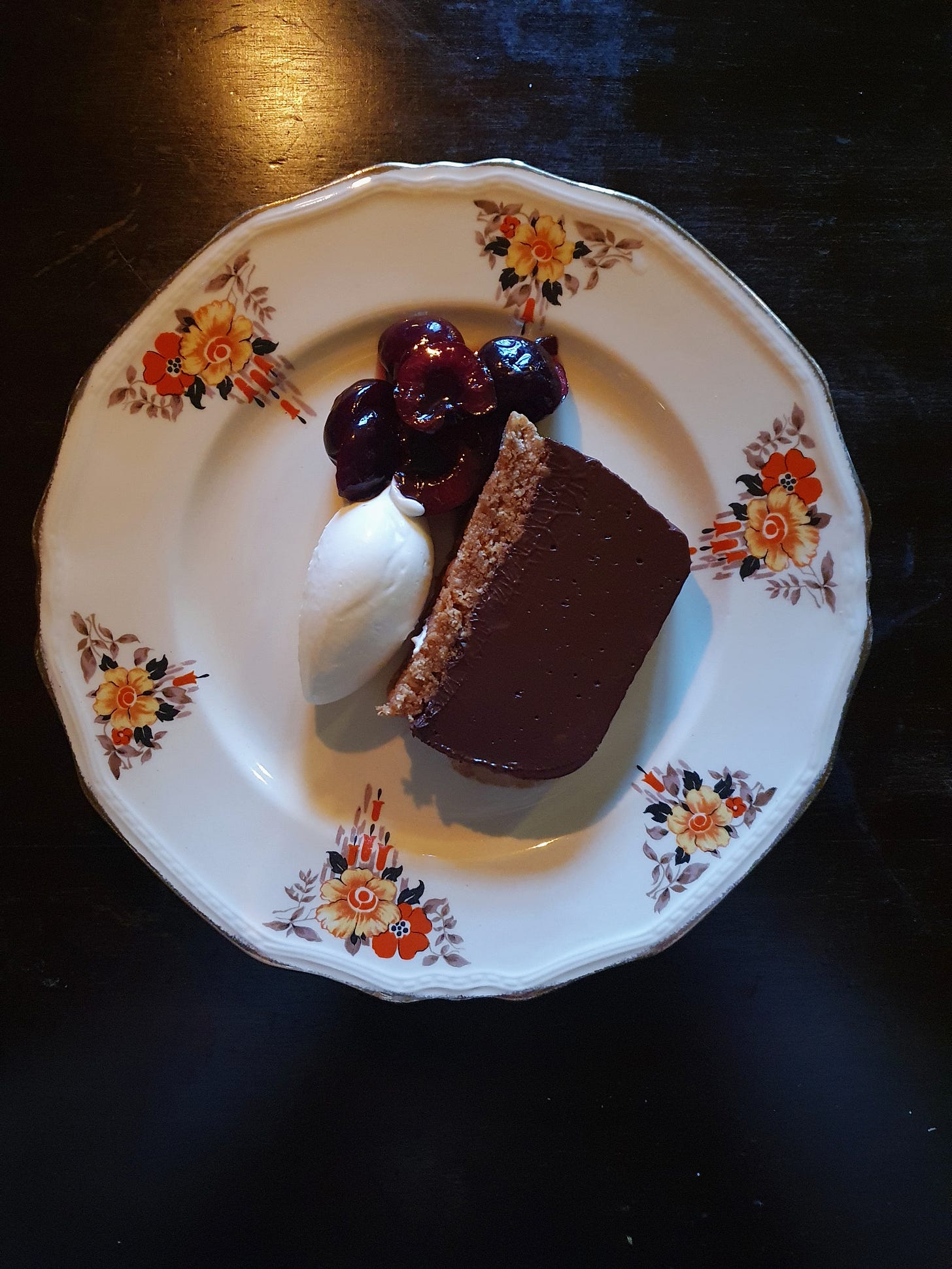 a slice of chocolate tart on a pretty plate decorated with flowers, also on the plate are some dark cherries and a quenellle of creme fraiche
