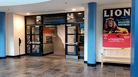 Museum building exterior, lobby with large sign "Steinbeck:, and entrance with statue of seated Steinbeck peeking out