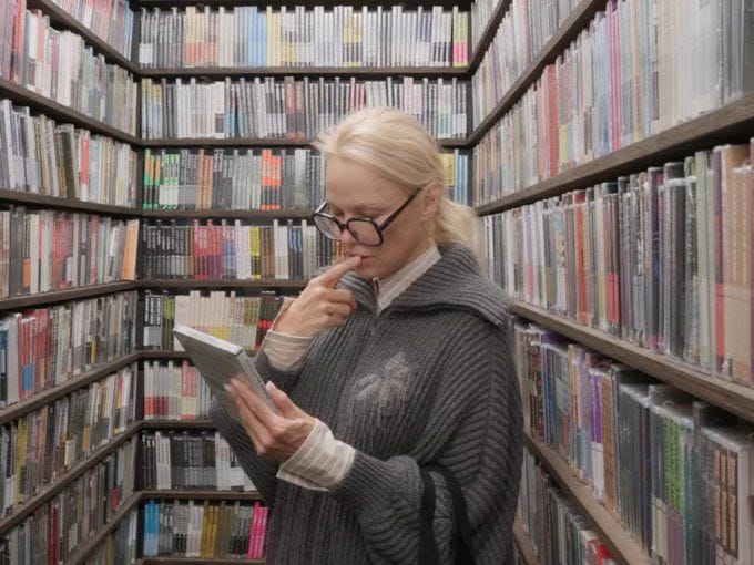 This may contain: a woman standing in front of a bookshelf looking at something on her tablet This may contain: a woman standing in front of a bookshelf looking at something on her tablet