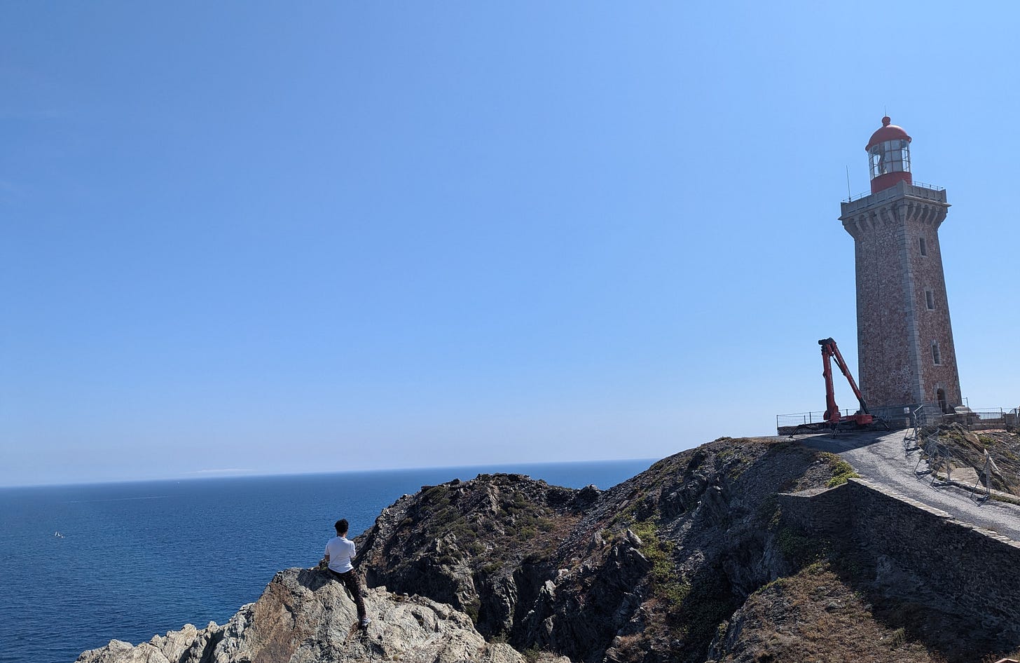 Lighthouse at Cap Bear, brick built with a red top housing the light. A woman sits on the cliffs admiring the view of the sea