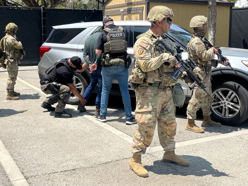 Full photo. A guy with a POLICE vest handcuffs a different person., alongside another guy in a plate carrier with his face fully covered. Next to them are three men in military fatigues carrying a rifle. One of them is wearing a neck gaiter covering his face.