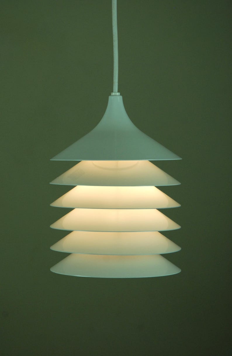 May include: White pendant light fixture with a layered, conical design. The light has a series of stacked, inverted cone-shaped shades, creating a tiered effect. The fixture hangs from a white cord against a muted green background. May include: White pendant light fixture with a layered, conical design. The light has a series of stacked, inverted cone-shaped shades, creating a tiered effect. The fixture hangs from a white cord against a muted green background.