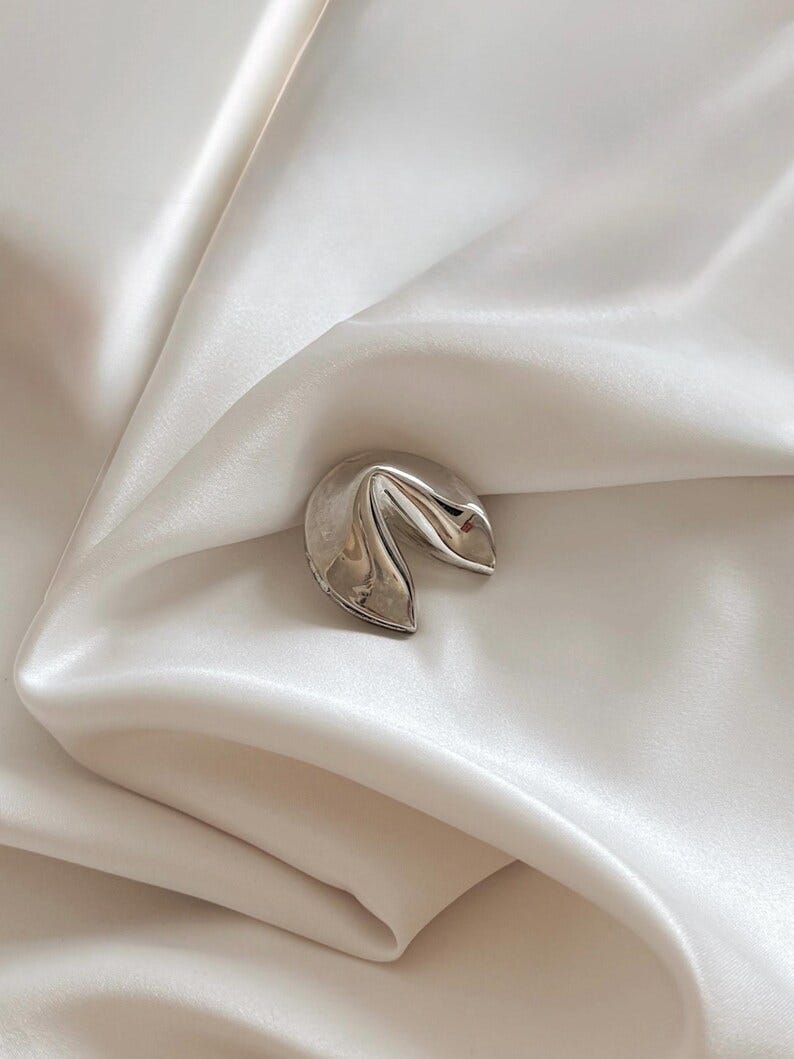 May include: A shiny silver fortune cookie charm rests on a white satin surface. The charm is highly reflective, with a smooth, curved shape. The background is a soft, off-white color, creating a contrast with the metallic charm.