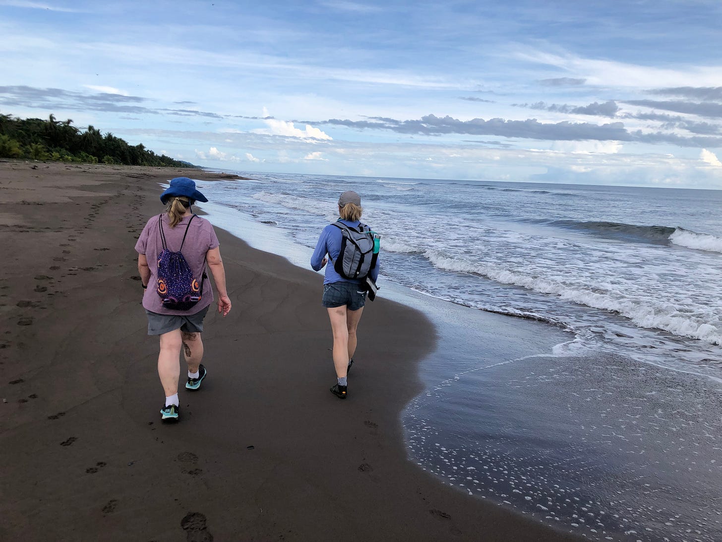 Two ladies walk along black sand on the beach with small backpacks on their backs.