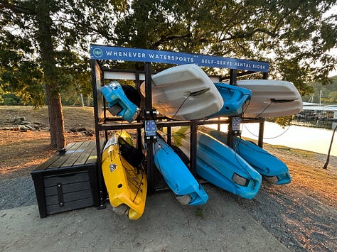 The left image shows a storage rack of eight blue, yellow, and gray kayaks for rent. The middle image shows a QR kayak checkout station, with a blue sign, a white sign letting Nicole know she can't sue Arkansas State Parks if she drowns, and a brown lockbox that holds oars and lifejackets. The final image is the sweet view of freedom: the front tip of a lime-green kayak is visible above a large lake of soft, deep blue waves.