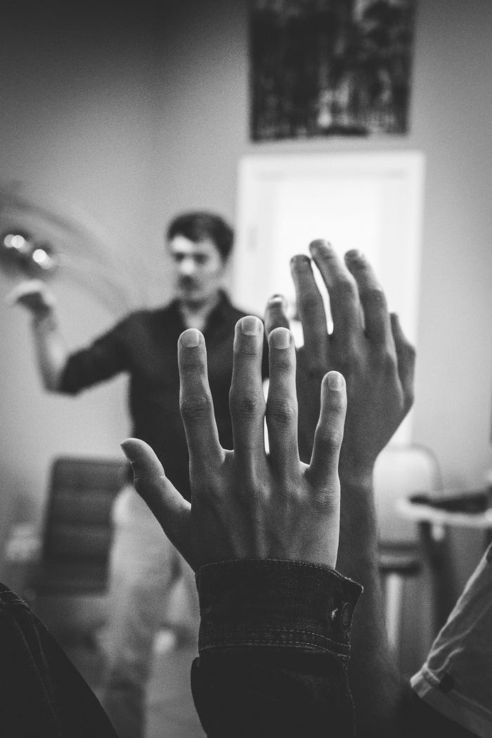 People raising their hand to ask questions as a man in the background is talking to a crowd. People raising their hand to ask questions as a man in the background is talking to a crowd.