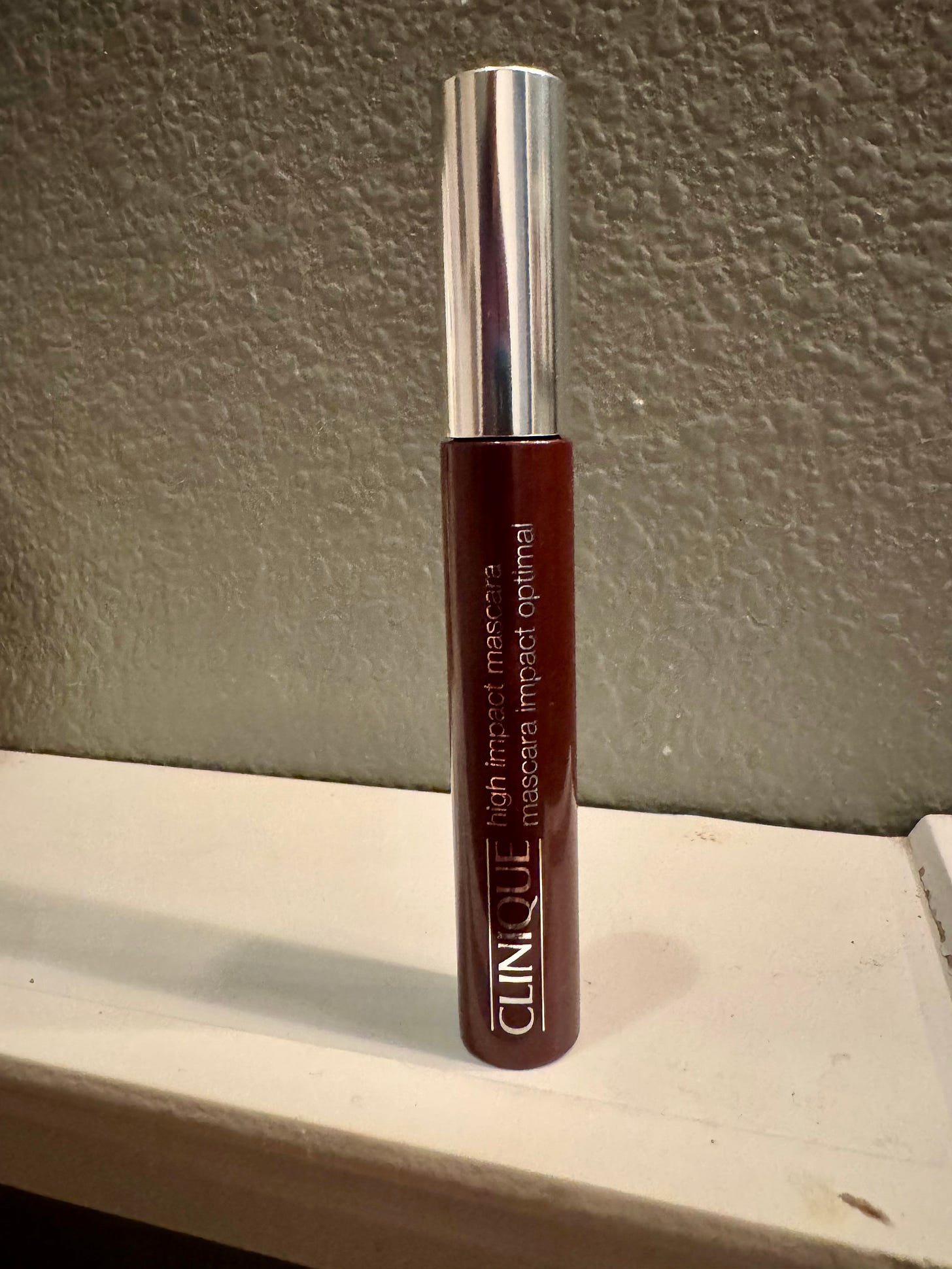 A single tube of Clinique High Impact Mascara with its iconic silver cap and dark brown-green body, standing upright on a white ledge against a textured gray wall background.