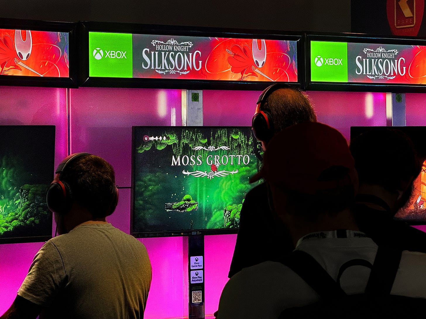Photo of gamers playing Silksong on monitor screens, set against a purple wall with backlit banners featuring character artwork.