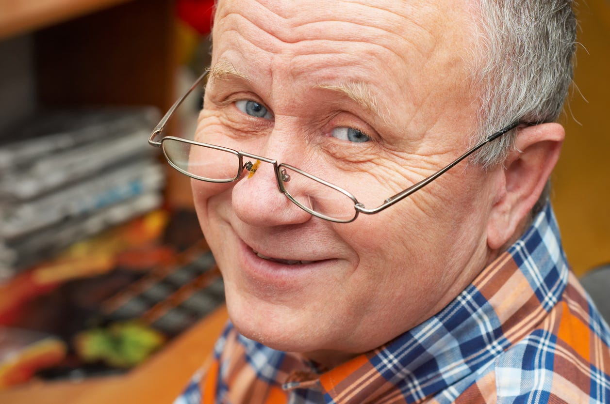 Tight facial shot of aging, smiling white man with half eye glasses on the tip of his nose, exposing bright blue eyes..