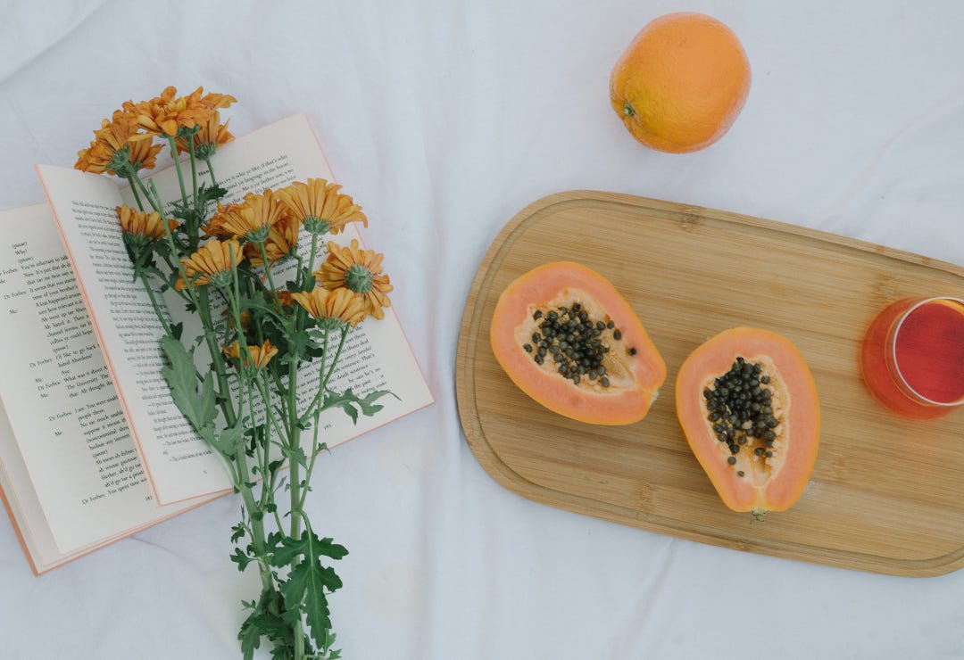 On a white cloth lays an open book with a small bouquet of orange marigold flowers in between the pages. Next to the book lies a wooden cutting board with a papaya cut open revealing the black seeds, a glass of orange tea, and a whole orange. 