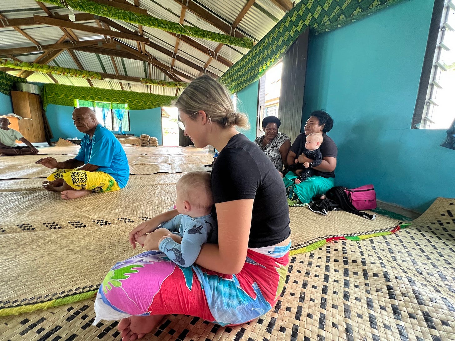 A woman sits on a straw mat with a baby on her lap. Several people sit cross-legged in the background. 