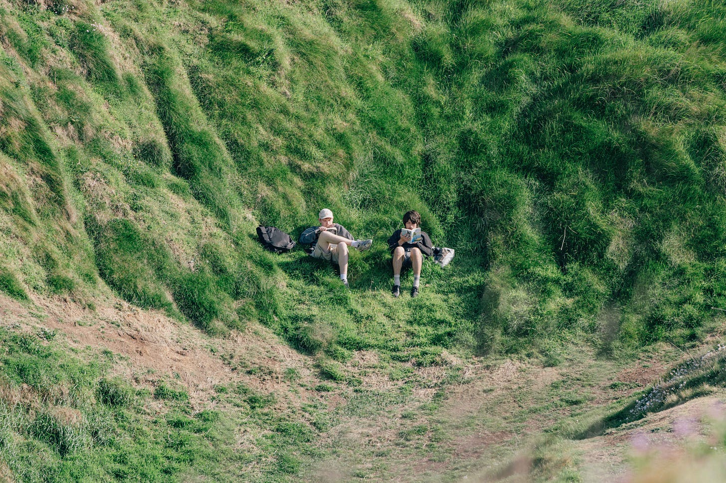 Two people lie back in a grassy hollow, reading and resting under soft afternoon light, framed by uneven cliffside grass. Two people lie back in a grassy hollow, reading and resting under soft afternoon light, framed by uneven cliffside grass.