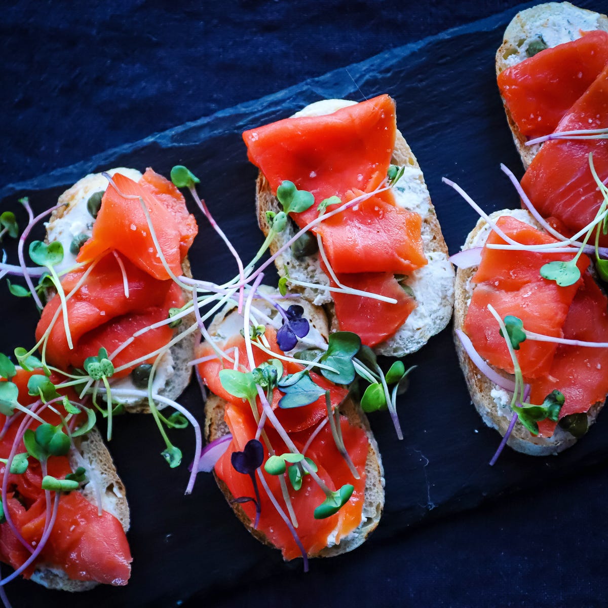 Small crostini topped with deep red, smoked salmon slices, cream cheese and some microgreens on a black platter.