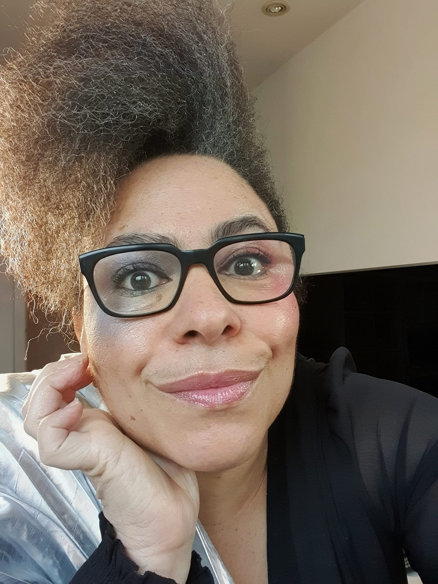 A woman wearing glasses looking at the camera. She has one cheek and eye made up with silver and the other with pink makeup.