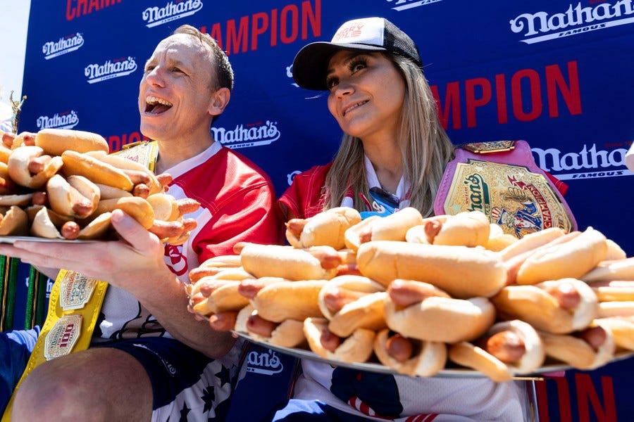 A man and a woman pose while holding up platters overflowing with dozens of hot dogs.
