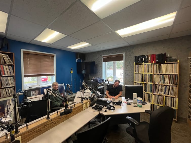 Two people inside a radio studio, sitting at a long table-desk combination. Two people inside a radio studio, sitting at a long table-desk combination.