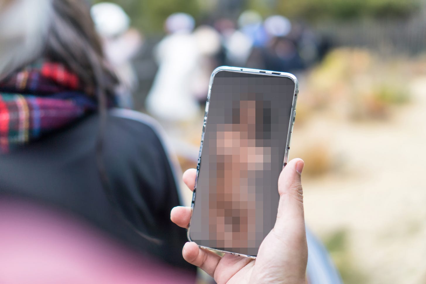 A hand holds a smartphone with a heavily pixelated image on its screen, a crowd of people blurred in the background. A hand holds a smartphone with a heavily pixelated image on its screen, a crowd of people blurred in the background.