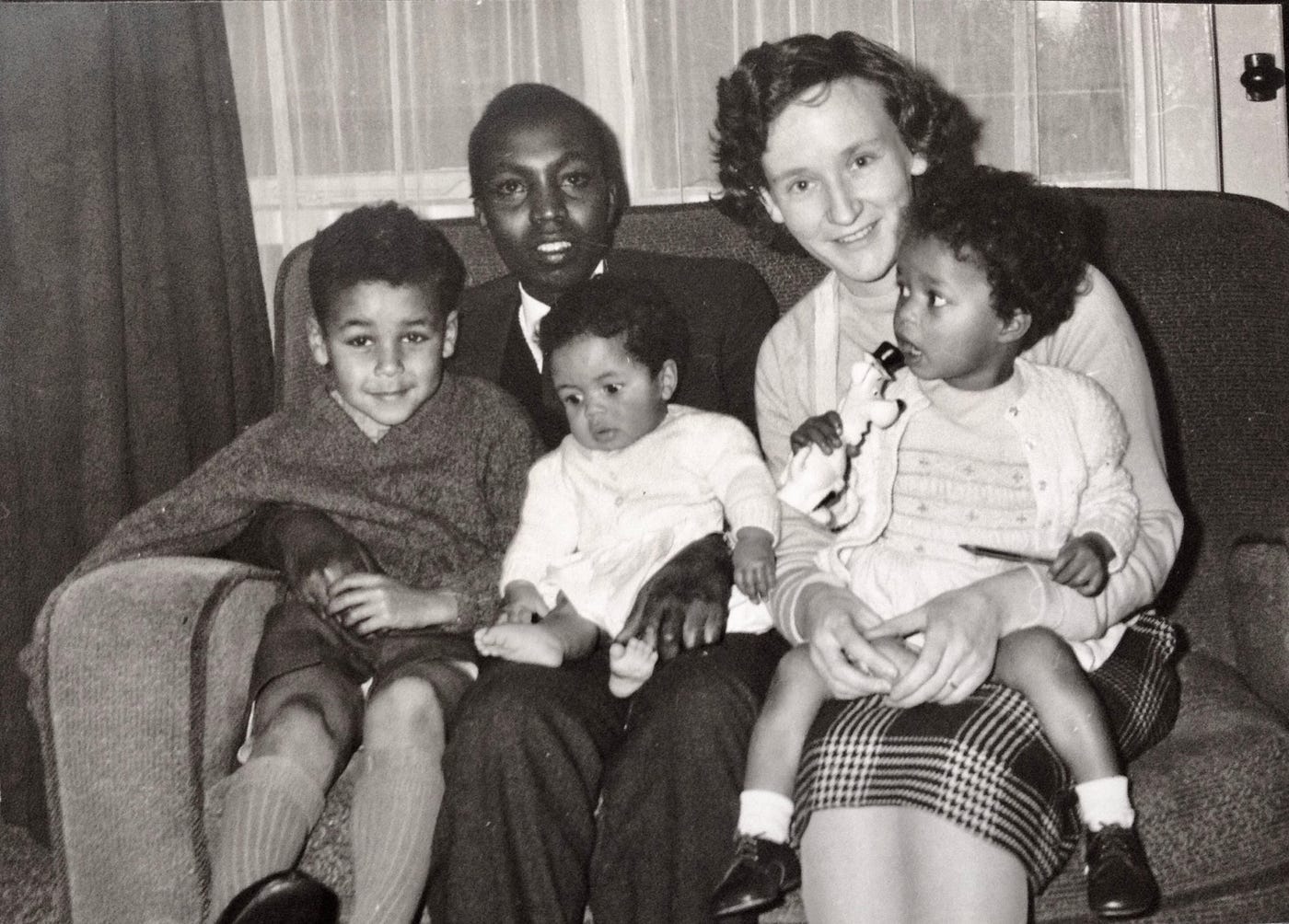 Old black & white photo of my Kenyan father next to my Scottish mother on a sofa with me age 3, and my 2 brothers, 8 and 18 months. Everyone smiles except the youngest who is looking at his toes, and me looking distracted.