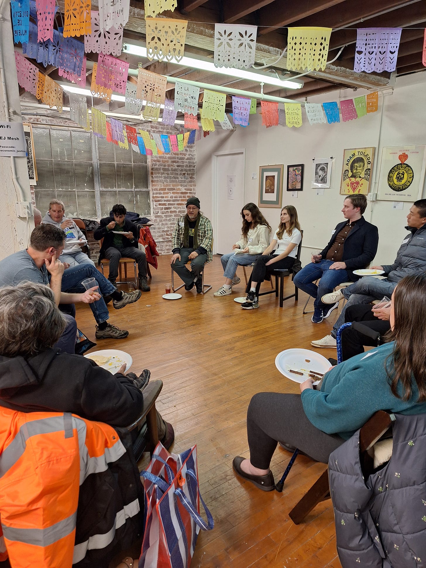 A circle of people sitting in chairs, with a wide diversity in ages, all listen to one person speaking while they eat. The room they sit in is a large, open space with multicolored paper decorations on the ceiling. A circle of people sitting in chairs, with a wide diversity in ages, all listen to one person speaking while they eat. The room they sit in is a large, open space with multicolored paper decorations on the ceiling.