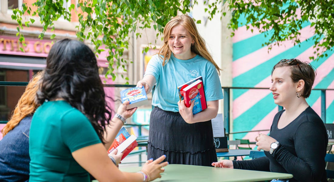 A woman handing out a book to a group of people sitting at a green table