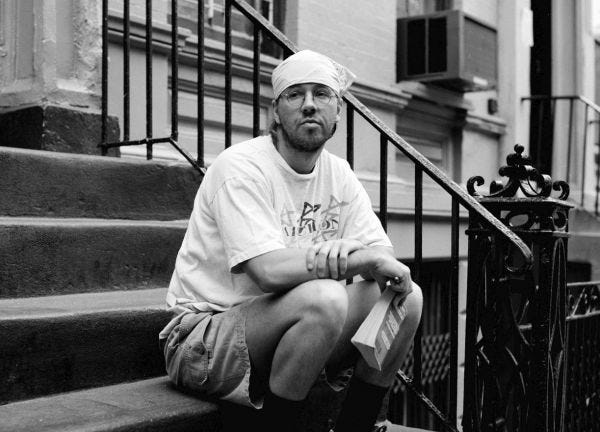 Escritor David Foster Wallace, numa foto em preto e branco, sentado em uma escada a frente de uma casa