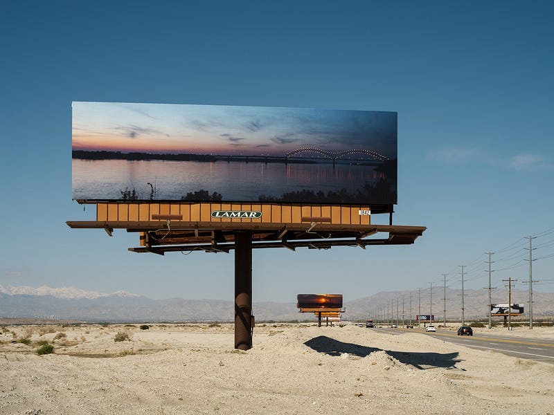 The sandy shoulder of a highway with rows of billboards on either side. In the foreground is a billboard covered by a sunset photo of a body of water, with a bridge off in the distance. The sandy shoulder of a highway with rows of billboards on either side. In the foreground is a billboard covered by a sunset photo of a body of water, with a bridge off in the distance.