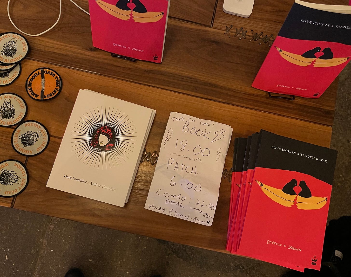An overhead view of part of the merchandise table. There are patches, Derrick's book "Love Ends in a Tandem Kayak," and Amber's book "Dark Sparkler." There is a small piece of paper with prices handwritten on it in pen.