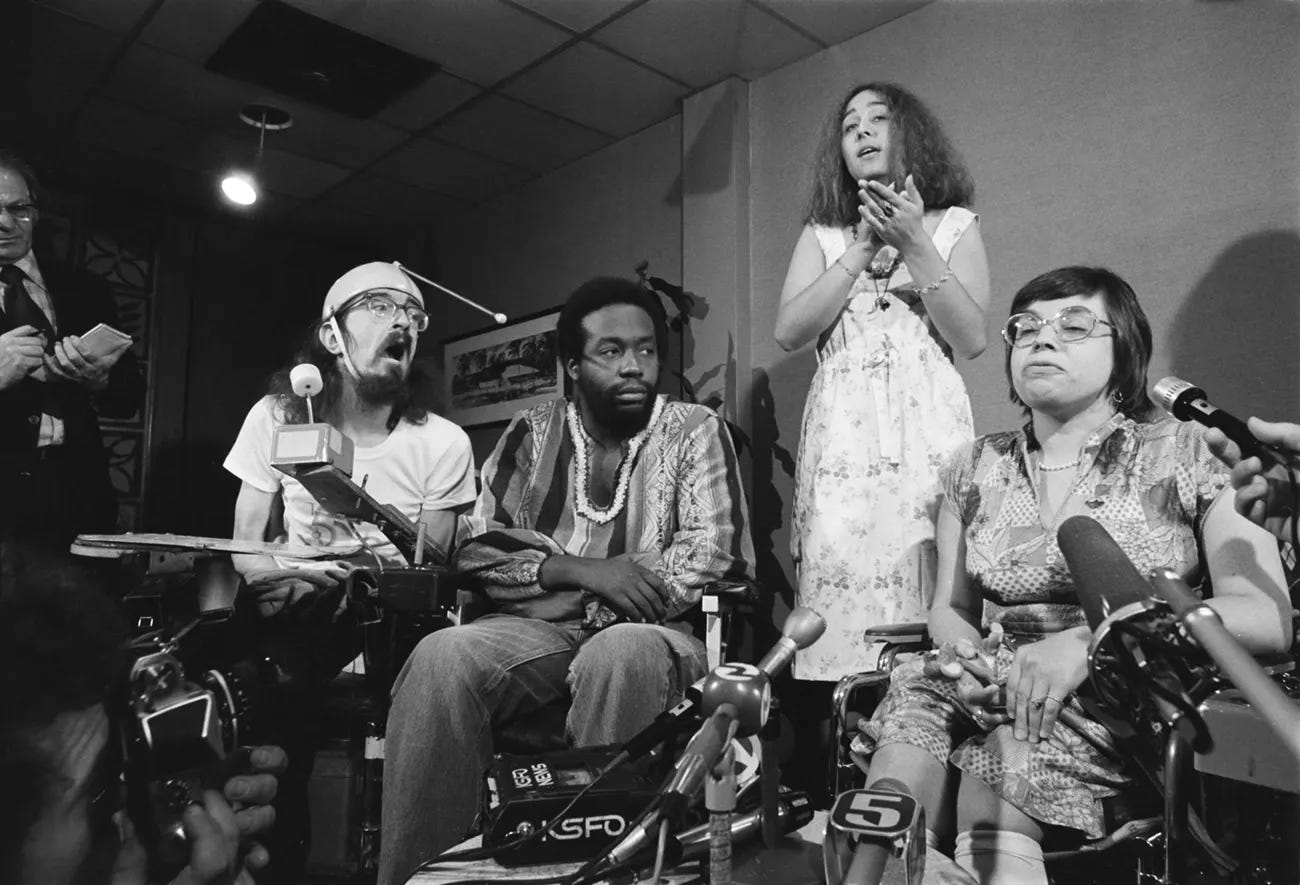 At a press conference, disability rights activists Hale Zukas, Ron Washington, and Judy Heumann sit in wheelchairs behind a row of microphones as reporters and cameras crowd around them. Behind them, Lynette Taylor interprets in American Sign Language.