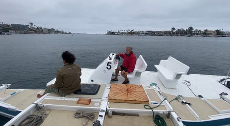 small voyaging canoe up on the sandy beaches on a gray chilly day. Our friend Kalani stuffing and trying to manage many bags and tools stuffed into a car seat. An uncle towing our boat with a side tie and a small engine boat used for paddling races.