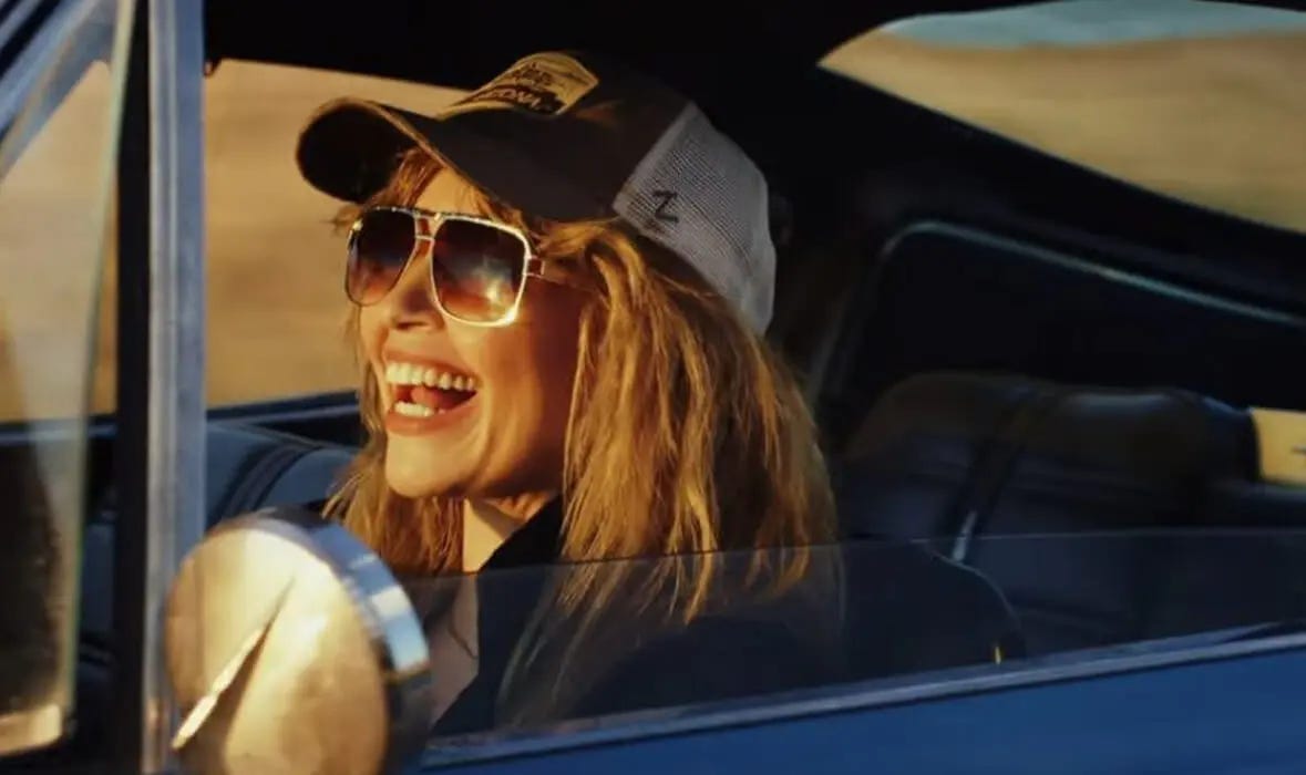 blond woman wearing a baseball cap laughs while driving a car
