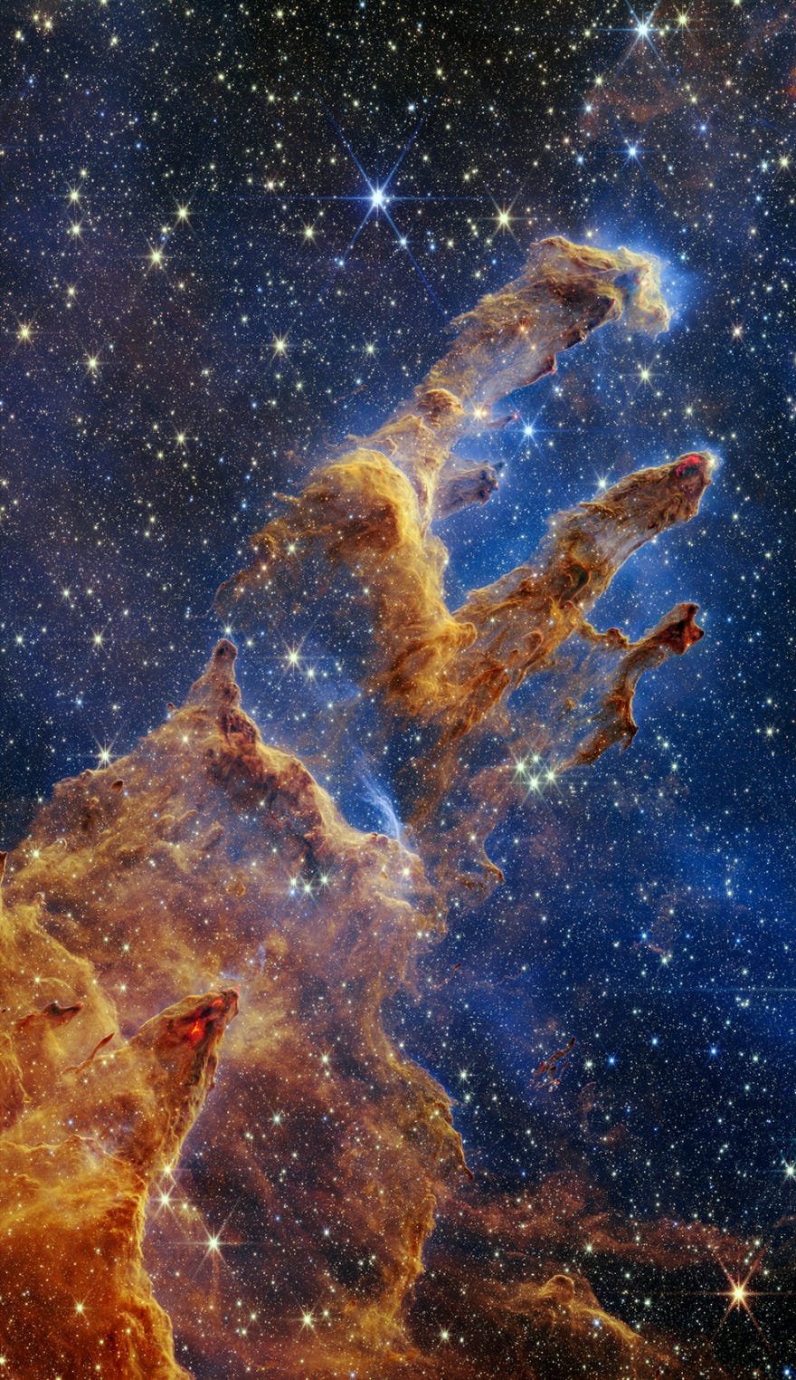 A large image of space shows a dark blue night sky with thousands of very bright stars of varying sizes. In the middle of the image is a brown and tan gaseous cloud that forms three roughly shaped pillars that extend upwards towards the upper right-hand corner of the picture. At the tips of several of the pillars are small, bright red spots that appear to be fire. A large image of space shows a dark blue night sky with thousands of very bright stars of varying sizes. In the middle of the image is a brown and tan gaseous cloud that forms three roughly shaped pillars that extend upwards towards the upper right-hand corner of the picture. At the tips of several of the pillars are small, bright red spots that appear to be fire.