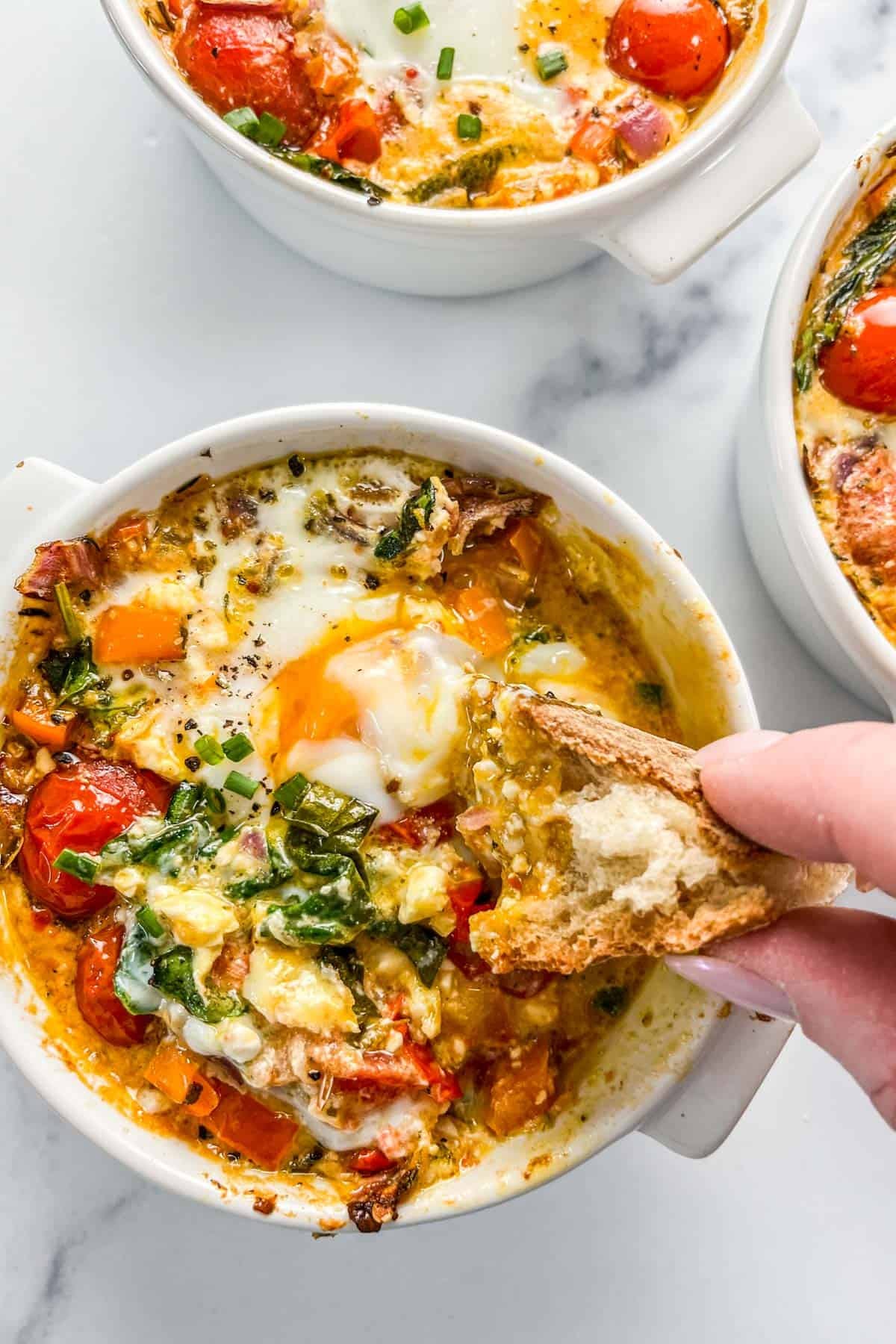 A piece of bread being scooped into a ramekin of baked feta eggs.