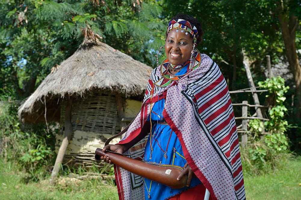 Jemimah Kutata, a Kenyan woman, stands, smiling. She is wearing traditional Maasai clothes and stands in front of a thatch-roof hut. Jemimah Kutata, a Kenyan woman, stands, smiling. She is wearing traditional Maasai clothes and stands in front of a thatch-roof hut.