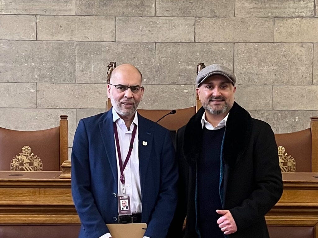 Ran away Akbar stands in a navy blue suit and white shirt next to Faraz Arian, who wears a grey hat and dark coat. They are standing in front of oak panelled pews and a grey brick wall.