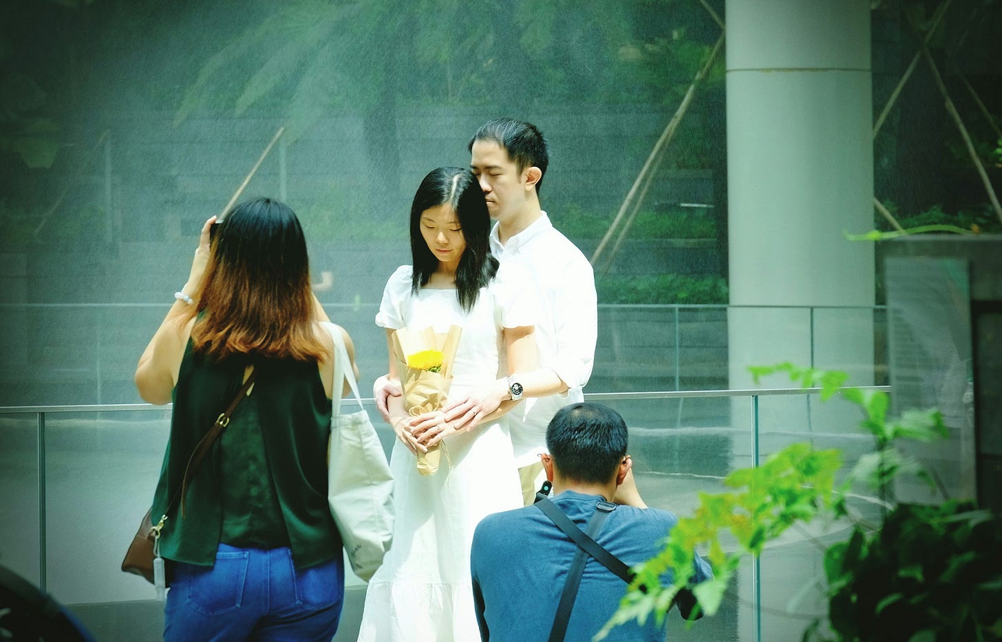 A young couple poses for a wedding-style photoshoot in Singapore, with photographers capturing the moment. The woman in a white dress holds a bouquet while the man stands closely behind her, highlighting the local creative industry. A young couple poses for a wedding-style photoshoot in Singapore, with photographers capturing the moment. The woman in a white dress holds a bouquet while the man stands closely behind her, highlighting the local creative industry.