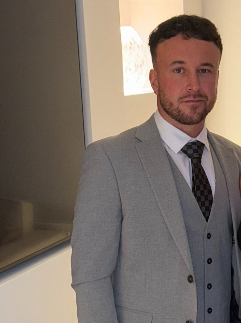 Man standing in front of a light with a white wall behind him. He has short dark hair, and short facial hair. He is wearing a light grey suit and dark coloured tie.