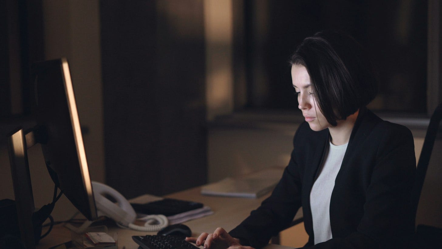 A woman in a dark blazer types at a desktop computer in a dim office after hours, with a desk phone and notebooks beside the monitor. A woman in a dark blazer types at a desktop computer in a dim office after hours, with a desk phone and notebooks beside the monitor.
