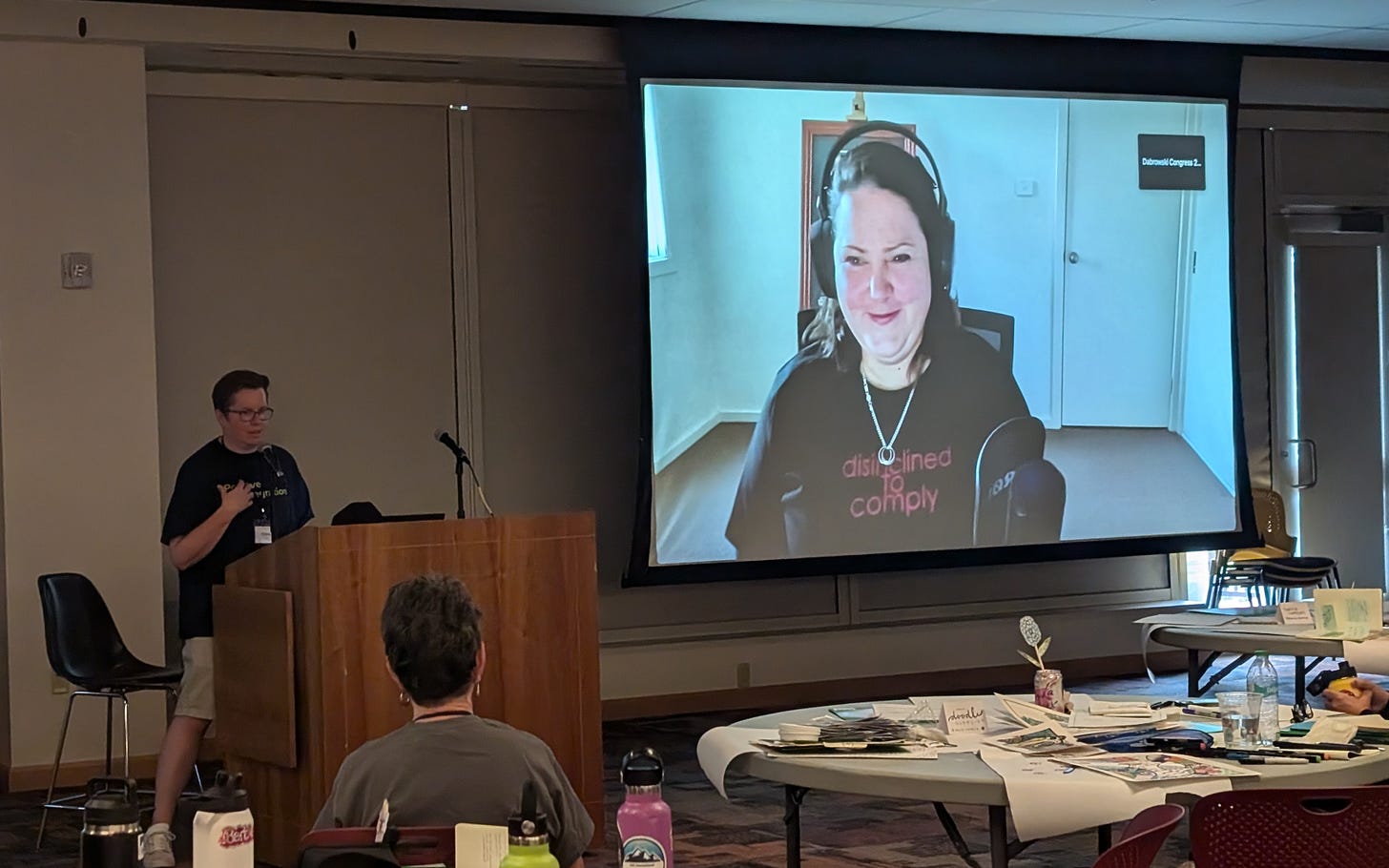Image description: Chris Wells at the podium, looking at Emma on screen, while Emma is watching Chris from the giant screen in the room. Linda Silverman is at a table in front, watching. 