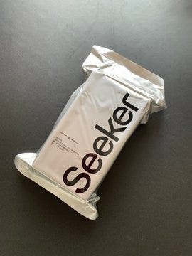 A white Solana Mobile Seeker device in its packaging, lying on a dark surface. The packaging has the word "Seeker" in large black letters and smaller text. A smartphone screen displaying the Solana wallet app, showing a balance of $0.00, tokens, and collectibles options.